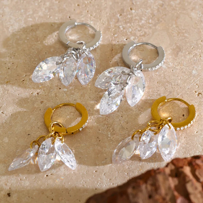 Four drop hoop earrings arranged on a beige stone surface, with two silver-tone and two gold-tone hoops, each featuring clear faceted teardrop crystal drops. Stainless steel, waterproof
