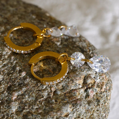 Gold hoop earrings with clear rhinestone accents and dangling clear faceted beads, displayed on a textured stone surface.