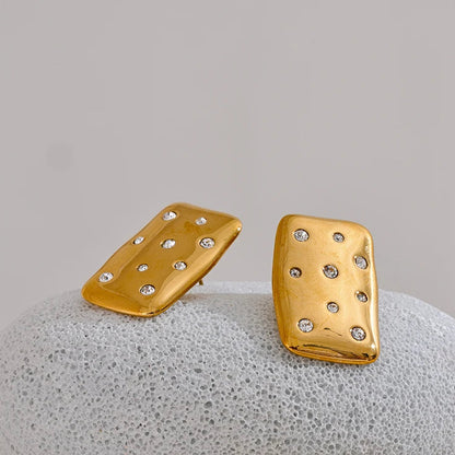 A pair of gold rectangular stud earrings with small clear rhinestones, displayed on a smooth gray stone against a light gray background. Stainless steel, waterproof