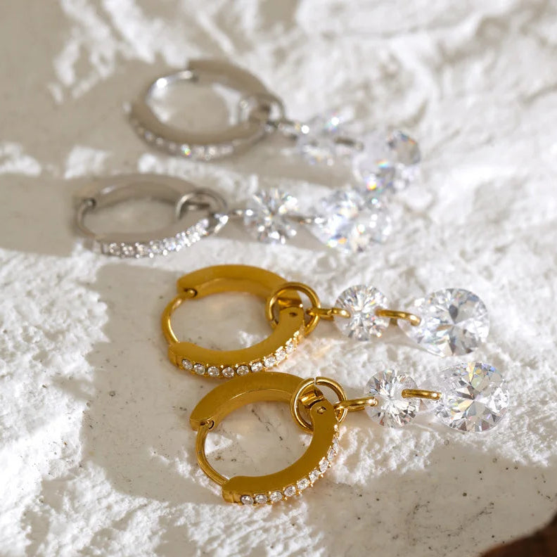 Two pairs of hoop earrings displayed on a textured white surface. One pair is gold with clear rhinestones, and the other is silver with clear rhinestones, both featuring dangling clear faceted crystal drops.