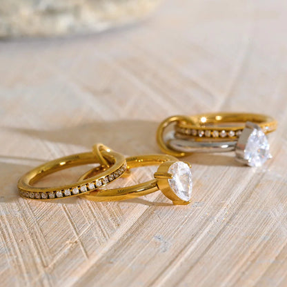 Two gold stacked rings with clear gemstones, including a pear-cut stone and a pavé band, displayed on a light wood surface.