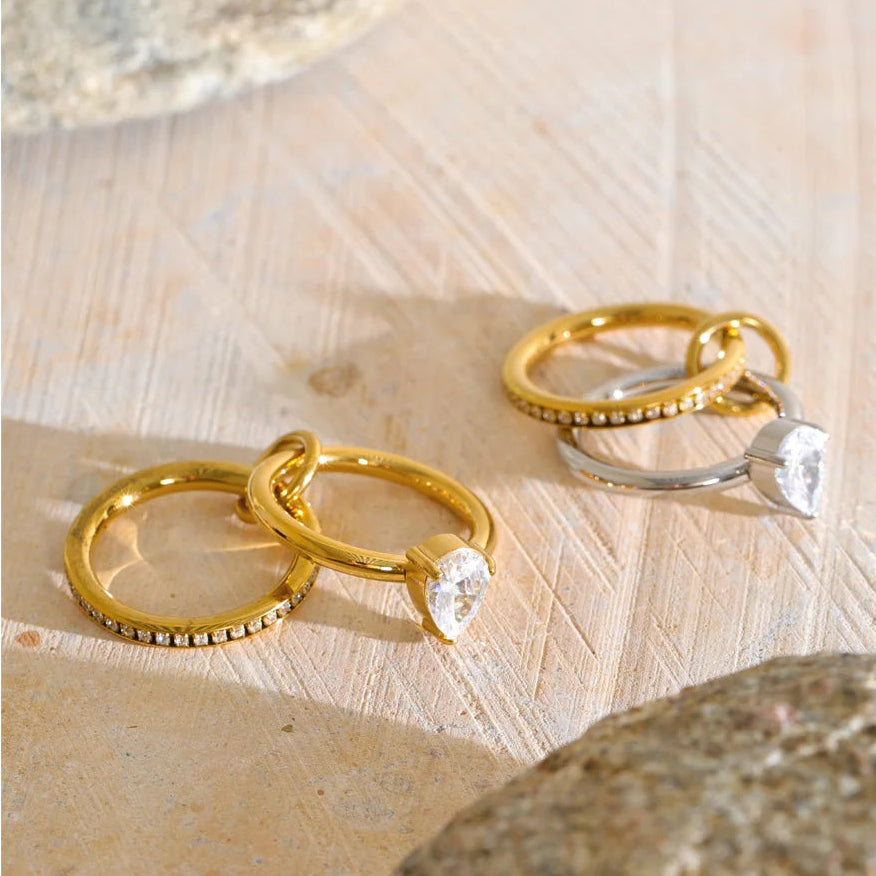 Two gold huggie rings with small pavé accents and heart-shaped clear gemstones, alongside a silver huggie ring with a larger clear heart gemstone, displayed on a textured beige stone surface.