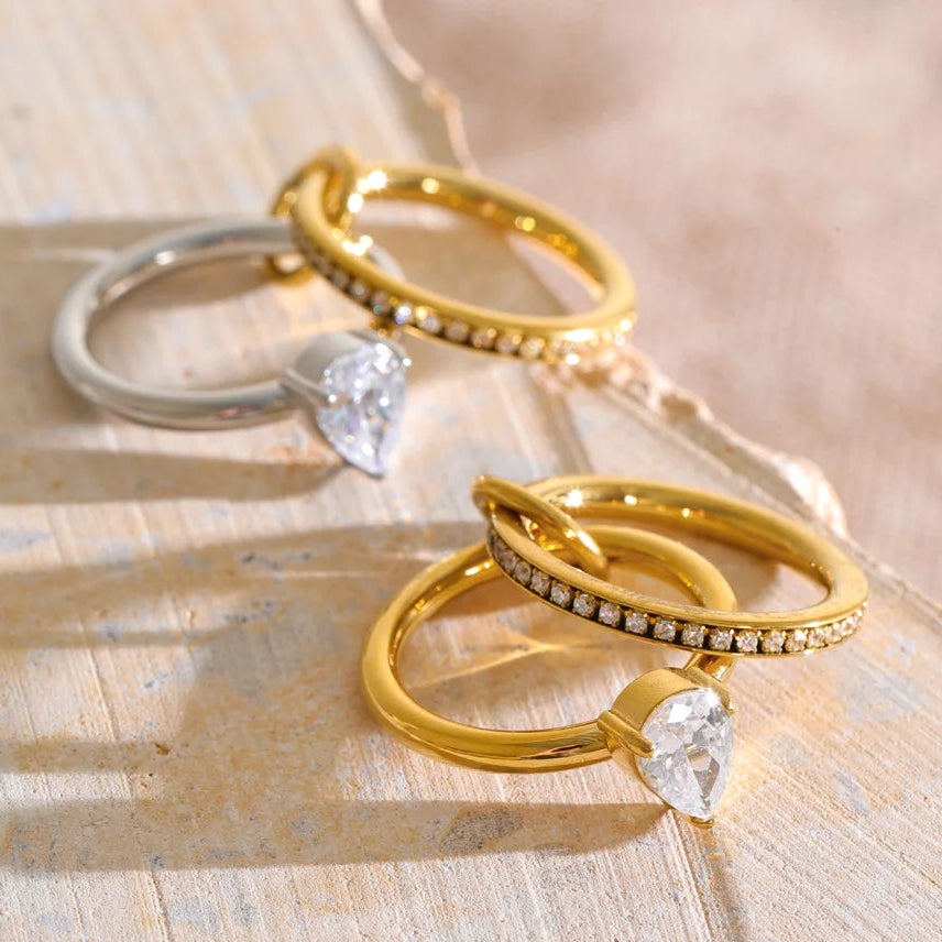 Three rings displayed on a light wood surface: two gold rings with small clear stones and one silver ring with a pear-cut clear stone.