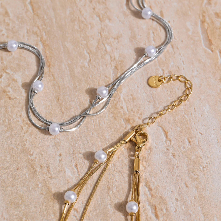 Two necklaces, one gold and one silver, with pearl beads on a beige stone surface.