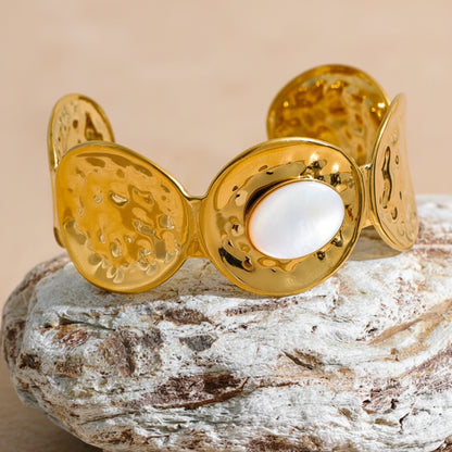 Gold bracelet with a central white stone on a textured surface with green foliage in the background