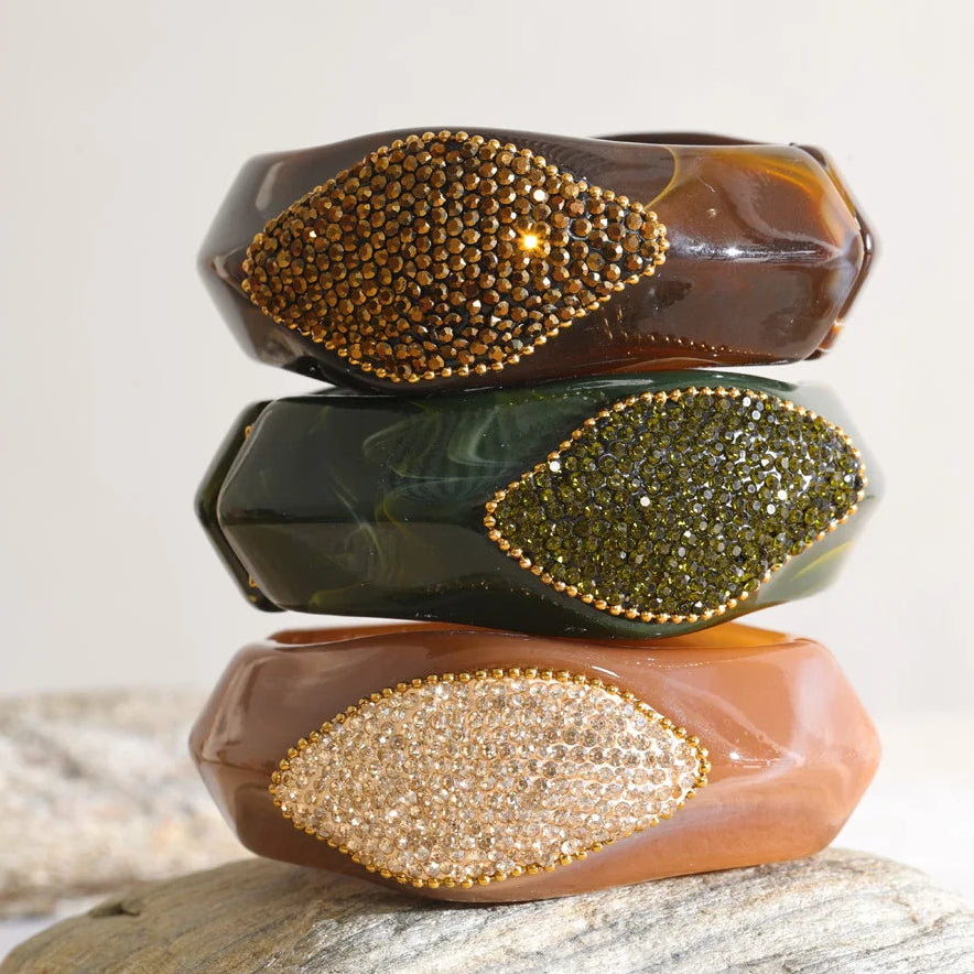 Three stacked translucent bangle bracelets with leaf-shaped crystal embellishments, displayed on a stone surface against a light neutral background.