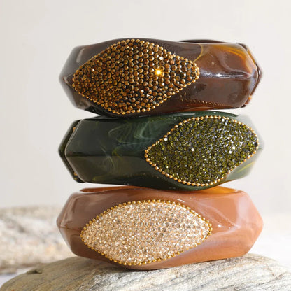 Three stacked translucent bangle bracelets with leaf-shaped crystal embellishments, displayed on a stone surface against a light neutral background.