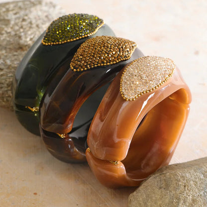 Three glossy tortoiseshell-style bangles with decorative leaf-shaped crystal embellishments in green, gold, and champagne tones, arranged on a light stone surface.