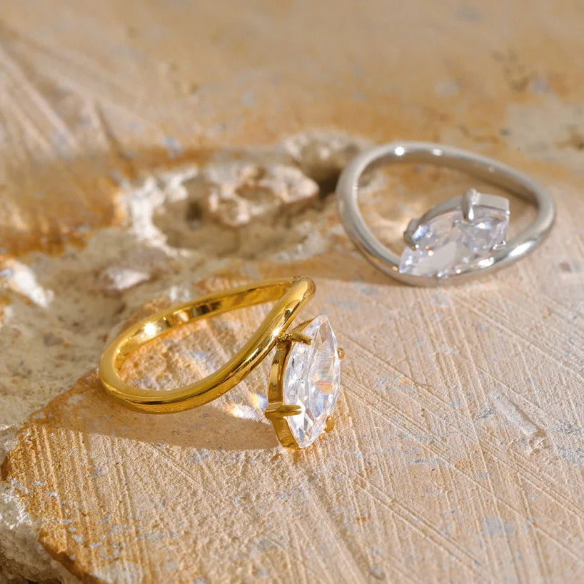 Two marquise-cut gemstone rings displayed on a textured beige stone surface. One ring features a polished yellow gold band, and the other features a silver-toned band.