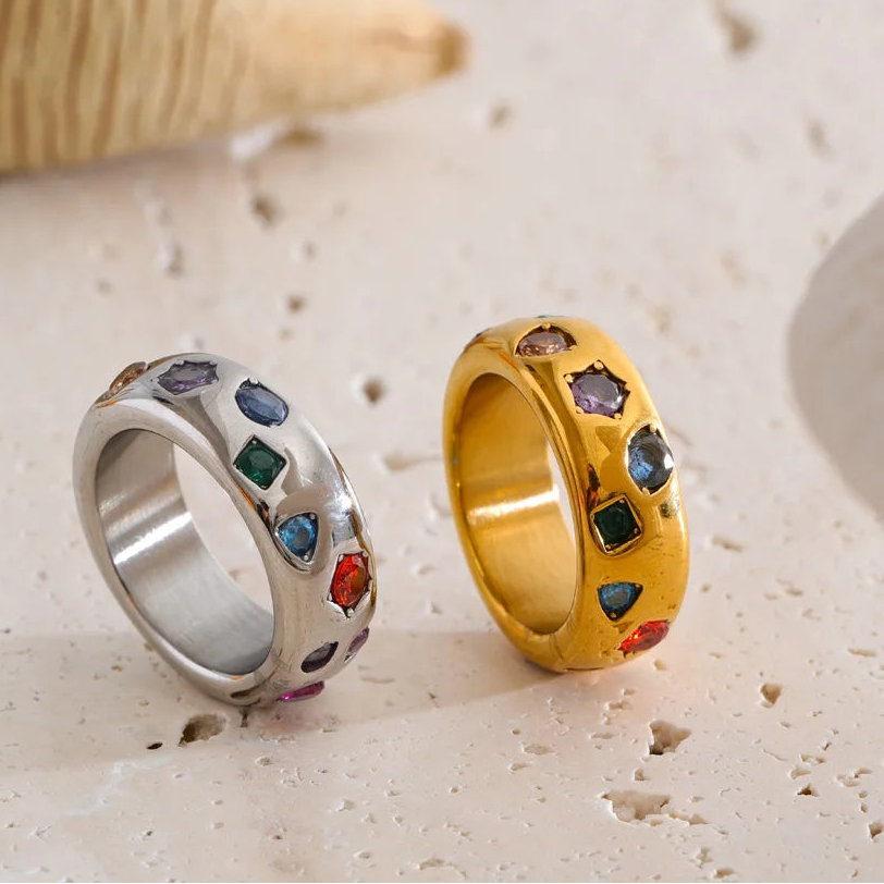 Two rings, one gold and one silver, with colorful gemstones on a sandy background.