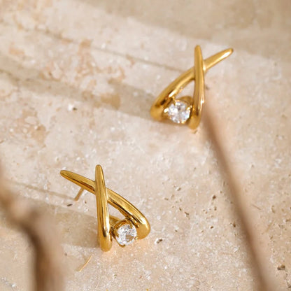 A pair of gold geometric stud earrings with a small clear gemstone, displayed on a textured beige stone surface with soft blurred branches in the foreground.