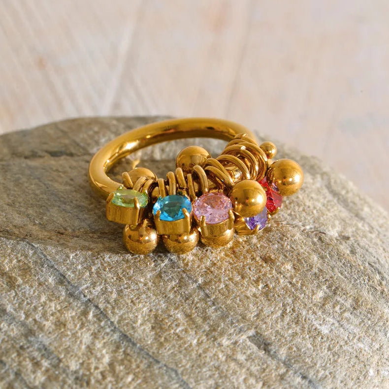 A gold ring with multiple small colorful gemstones and metallic bead details, resting on a textured stone surface.