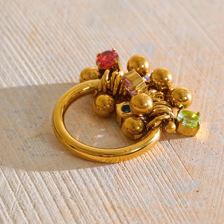 A gold-toned ring with a cluster of small gold beads and colorful gemstone accents, photographed on a textured light beige surface.