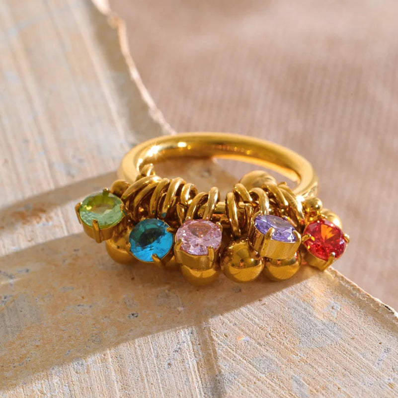 A gold ring with multiple colorful gemstones in green, blue, pink, purple, and red, resting on a textured beige surface.