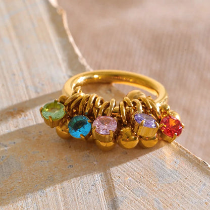 A gold ring with multiple colorful gemstones in green, blue, pink, purple, and red, resting on a textured beige surface.