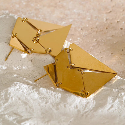 A pair of gold geometric dangle earrings with a faceted, multi-sided triangular design, displayed on a textured beige surface.