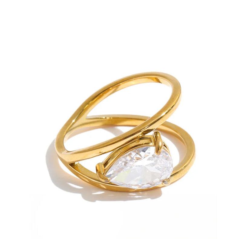 A gold ring with a twisted open-band design and a large teardrop-shaped clear gemstone, shown on a white background with a black script logo below.