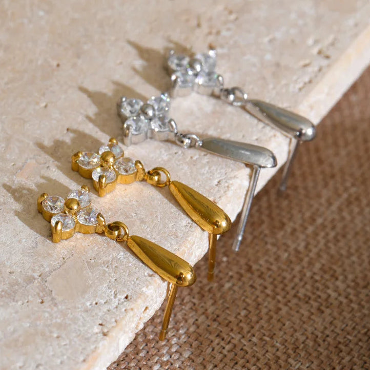 Four drop earrings displayed on a stone slab, showing both gold and silver metal finishes with clear crystal flower details and teardrop-shaped drops. Stainless steel