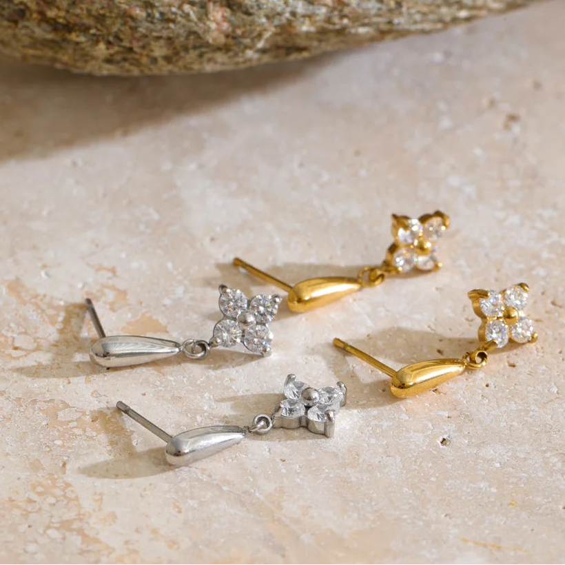 Four dangle earrings displayed on a light stone surface, including two gold and two silver pairs. Each earring features a small flower cluster with clear stones and a teardrop-shaped dangling element.