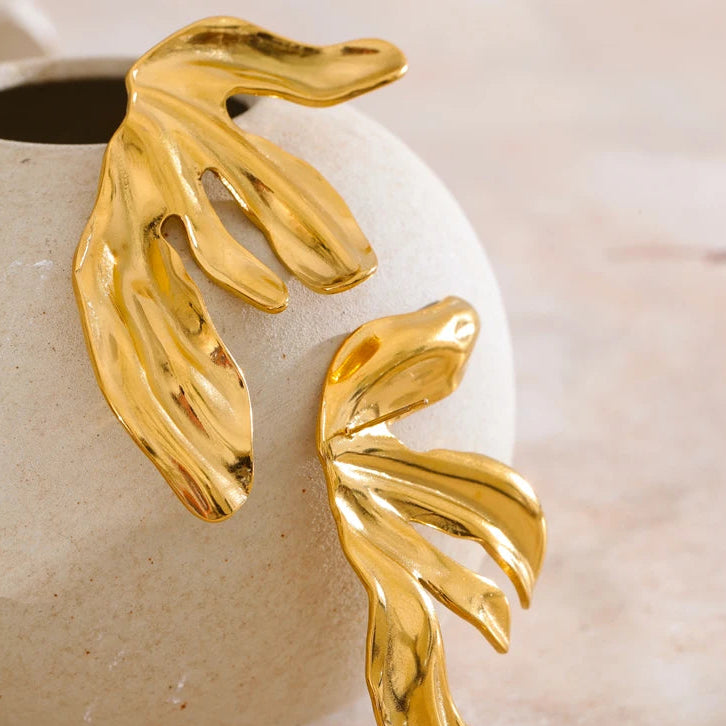 Gold leaf-shaped earrings on a beige ceramic surface