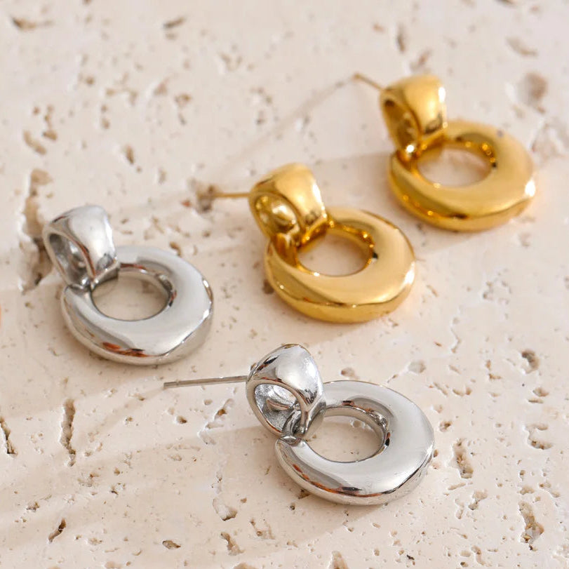 Three small ring-shaped drop earrings in polished silver and gold finishes, arranged on a light beige textured stone surface with a few brown stones partially visible at the edges.