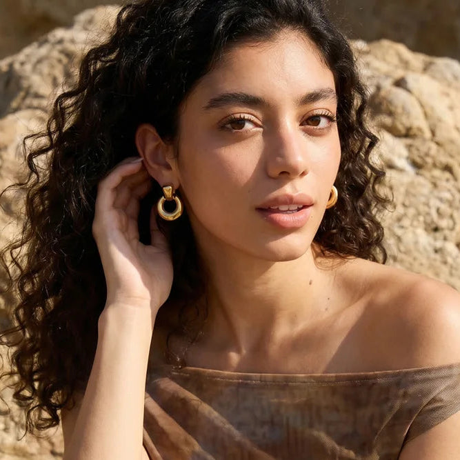 A woman with curly dark hair sits outdoors against large tan rocks, wearing a sheer off-the-shoulder top in a brown and beige tie-dye pattern and gold hoop earrings.