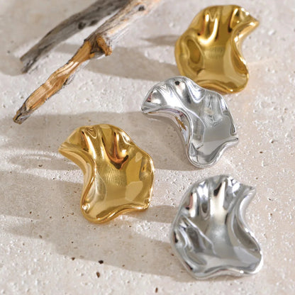 Four abstract, wavy metal jewelry pieces in polished gold and silver finishes, arranged on a textured beige stone surface with a piece of driftwood.