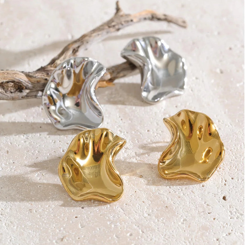 Four abstract sculptural earrings in polished gold and silver finishes, arranged on a light beige textured surface with a piece of driftwood.