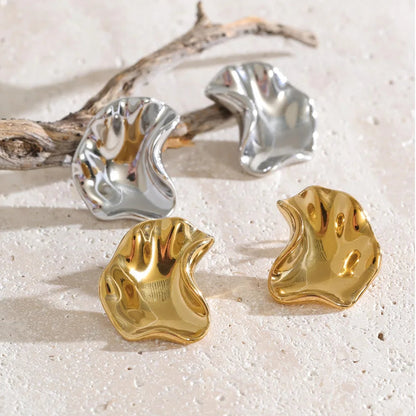 Four abstract sculptural earrings in polished gold and silver finishes, arranged on a light beige textured surface with a piece of driftwood.