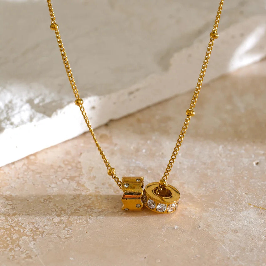 A gold chain necklace with two small cylindrical charms, one plain gold and one decorated with tiny clear stones, displayed on a textured beige stone surface.