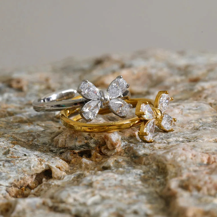 Two rings with butterfly-shaped clear gemstone settings, one in silver and one in yellow gold, displayed on a textured stone surface.