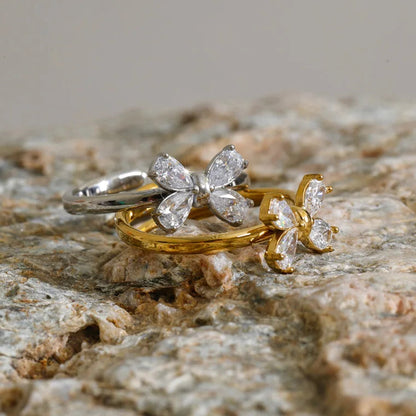 Two rings with butterfly-shaped clear gemstone settings, one in silver and one in yellow gold, displayed on a textured stone surface.